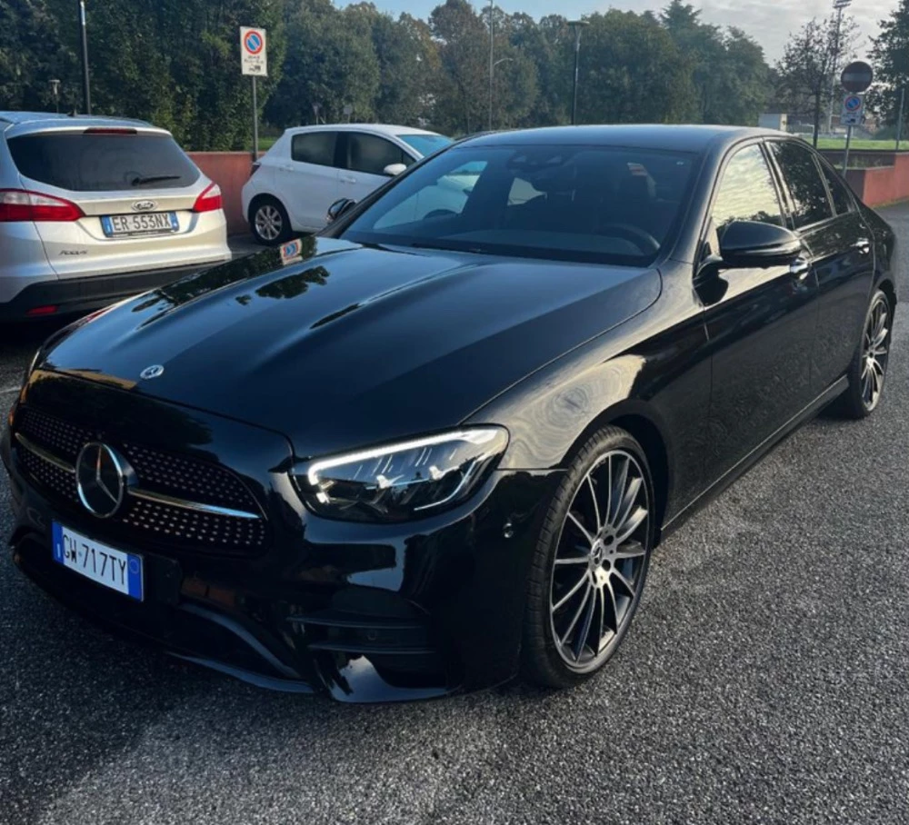 Mercedes Classe E nera con cerchi in lega e fari LED accesi, parcheggiata su asfalto tra altre auto e alberi